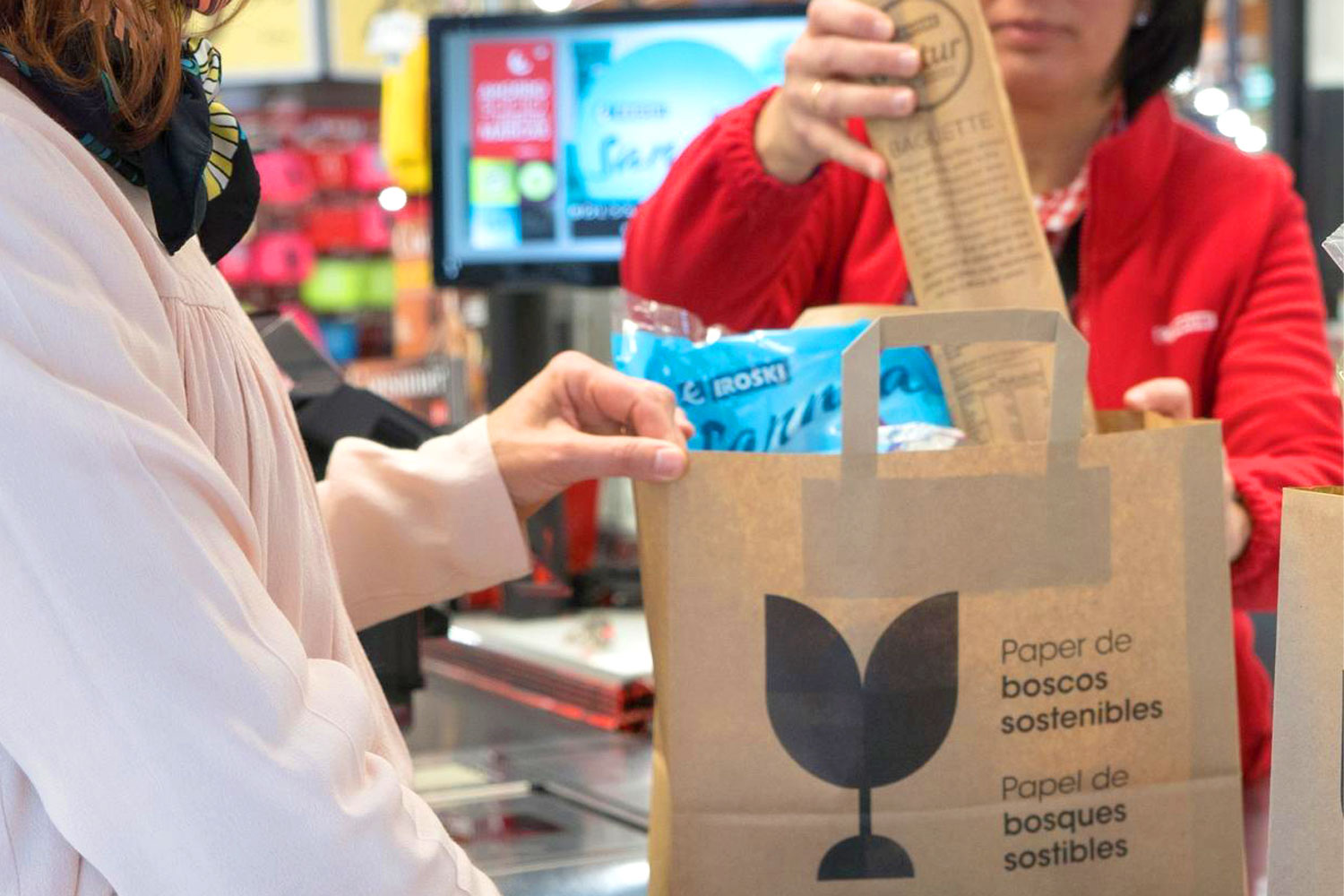 Client en caixa de supermercat rebent compra en bossa de paper sostenible mentre una empleada introdueix productes.