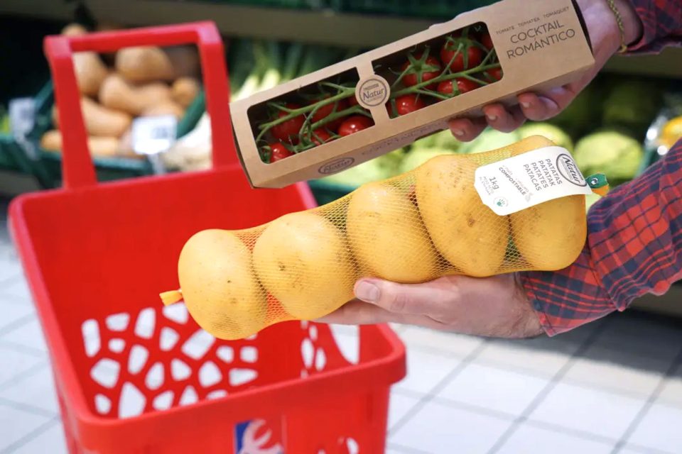Persona sostenint tomàquets en envàs de cartó i patates en malla compostable al costat de cistella de compra.