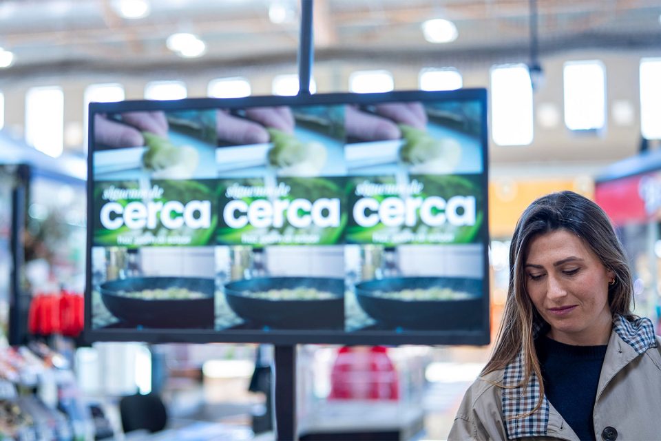 Dona en supermercat al costat de pantalla digital promocional amb missatges de proximitat i cuina preparada.