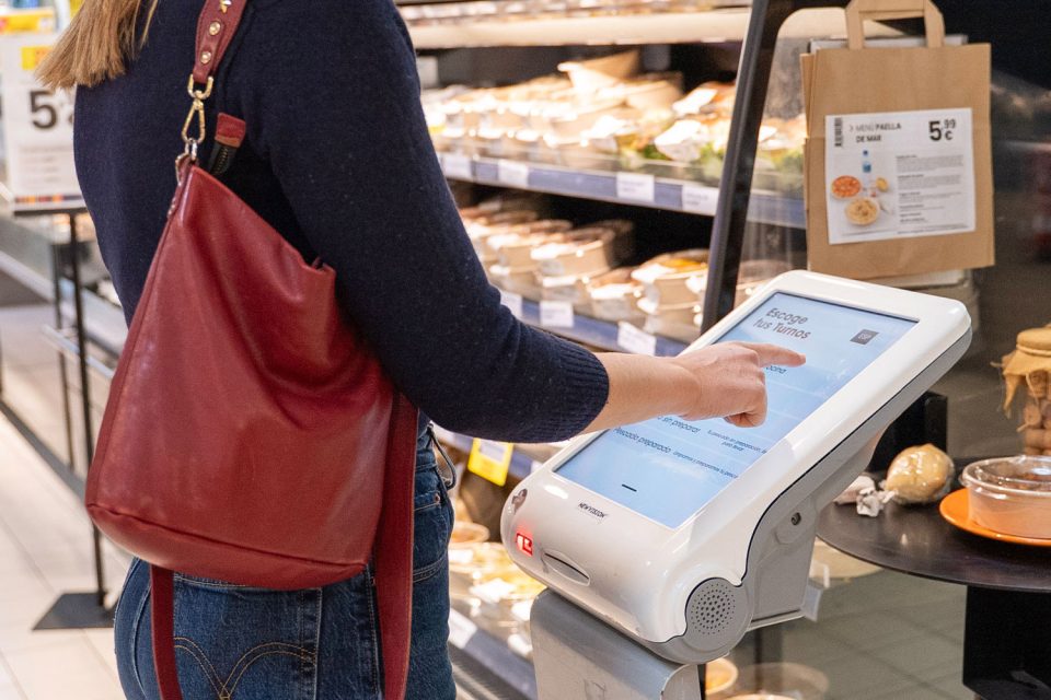 Clienta utilitza pantalla tàctil d'autoservei per a seleccionar torn en la secció de menjar preparat del supermercat.