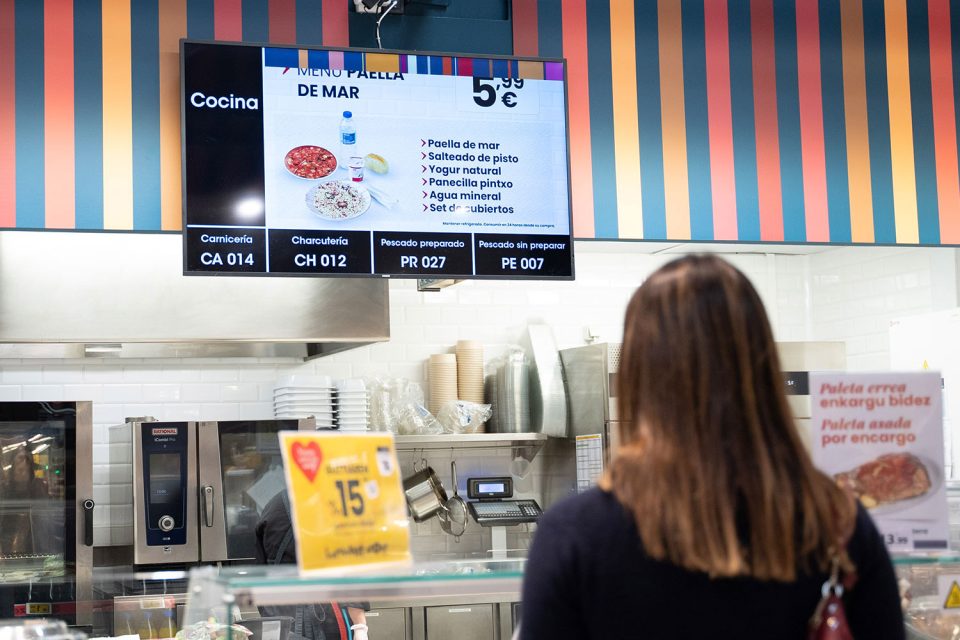 Clienta observa pantalla informativa amb menús i números de torn en la secció de frescos del supermercat.